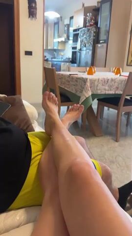 The video begins with a close-up shot of a person's legs and feet resting on a sofa, showing the knee and lower leg. The person is wearing yellow shorts and a black shirt. The camera then pans to the right, revealing a living room with a dining table and chairs in the background. A coffee table is visible with various items on it, including a phone, a bowl of potpourri, and glasses. In the distance, a television is playing a game show with a man as a guest, and a pet bed is on the floor in front of the television. The room is bright, and sunlight is visible through the windows.