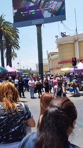 The video depicts a bustling city street filled with people, street vendors, and shops. 

The foreground shows several pedestrians, with one woman having long brown hair, partially obscuring the view. People are walking in both directions across a street. There are stalls set up with tents, displaying items for sale. A vendor is seated with a small cart selling Chicha, a drink. A street sign with the words “Santa Isabel” is visible. A clothing stall with items on hangers is set up.

The background contains palm trees and a large billboard advertising various companies. The sky is clear and blue, indicating a sunny day. 

The general atmosphere seems lively and crowded, typical of a busy street market.