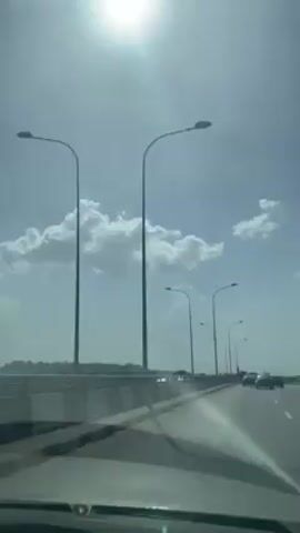 A vehicle travels down a freeway on a sunny day. The sky is light blue with scattered white clouds. Streetlights line the side of the road, and other vehicles can be seen in both directions. In the distance, a cell tower and a large building under construction are visible. The vehicle's windshield has some glare from the sun.