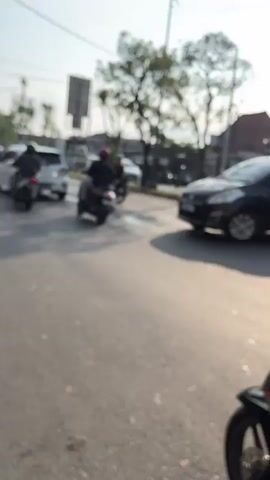 The video captures a street scene with several motorcycles and cars. In the foreground, two individuals are seated on a scooter. The person in front is wearing a red helmet and a dark long-sleeved shirt with patterned shorts. The person behind them is wearing a dark shirt and camouflage-patterned shorts. The scooter is black with red accents. To the left, another motorcycle with a rider in a black helmet and light brown jacket is visible. Further back, a white car is parked, and another motorcycle is moving. In the background, trees and utility poles line the street. The lighting suggests it is daytime. The video appears to be shot from a moving perspective, possibly from another motorcycle.