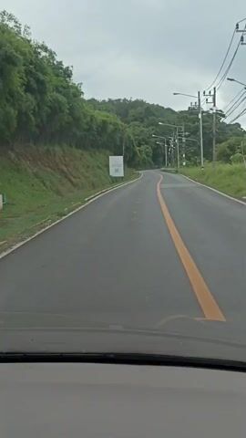 The video shows a view from a vehicle moving along a paved road. The road has a solid yellow line down the center and a white dashed line on the right side. To the left of the road, there is a grassy slope leading up to a densely wooded hill. To the right of the road, there are more trees and vegetation, along with a series of utility poles carrying power lines and streetlights. The sky is overcast and appears to be cloudy. There is a white sign with a QR code on it positioned on the left side of the road. The perspective suggests the video is taken from the driver's or passenger's seat of a car.
