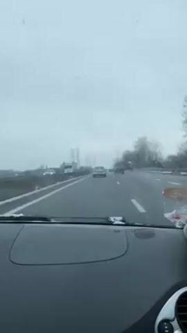 The video shows a driver's perspective while driving on a highway in snowy weather. The sky is overcast and snow is visible on the sides of the road. Cars are traveling in the same direction as the driver. Electrical towers are visible in the distance. The dashboard of the car is visible in the foreground. There is a phone mounted to the dashboard on the left side of the screen. The car's windshield wipers are on.