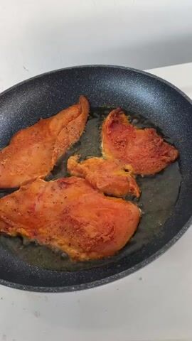 The video shows a close-up of raw chicken seasoned with a red spice blend being cooked in a non-stick skillet with oil. The chicken pieces are orange-red in color and appear to be sizzling in the hot oil. The camera then pans to a clear plastic container filled with more seasoned chicken. In the background, a white surface, possibly a countertop or stovetop, is visible. The video transitions to a wider shot of a table with various items, including a bottle of olive oil, a small cup with a tea bag, a small carton of milk, and some silver packets. The lighting suggests an indoor environment, possibly a kitchen or dining area.