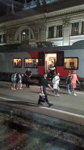 The video shows a train at a station at night. People are seen disembarking from the train. A few people are standing on the platform near the train. The train is gray and red with windows that are lit up from the inside. The building behind the train appears to be a train station with ornate architecture. The platform is concrete and there are train tracks visible in the foreground. The scene is dimly lit, indicating it is nighttime.