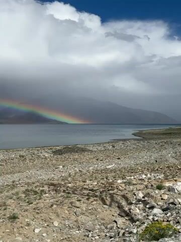 The video pans across a scenic landscape with a lake and mountains. A rainbow is visible in the sky, suggesting recent or ongoing rain. The foreground is rocky and sparsely vegetated. A motorcycle is parked on the left side of the frame in the later part of the video. The sky is partly cloudy with dramatic storm clouds.