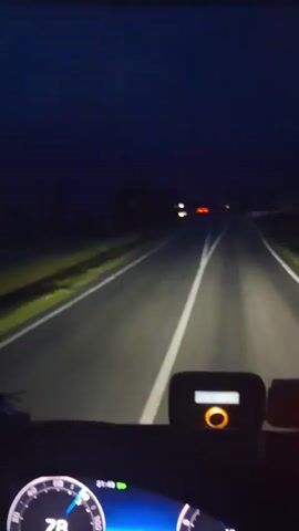 The video shows a first-person perspective from inside a vehicle driving at night. The view is through the windshield, focused on a two-lane asphalt road illuminated by the vehicle's headlights. White solid and dashed lane markings are clearly visible on the road surface, guiding the path ahead. The sky is dark, indicating nighttime. In the distance, the headlights and taillights of other vehicles can be seen, appearing as blurred points of light.

On the vehicle's dashboard, visible in the foreground, there are several objects. On the bottom left, a portion of a speedometer or gauge cluster is visible, showing numerical values and green indicator lights. In the center of the dashboard, a rectangular device with an orange ring and a small display screen is present. To its right, another device with a prominent flashing red light is visible. The immediate surroundings outside the road, such as the shoulders and distant landscape, are mostly dark or dimly lit by the vehicle's lights. The weather appears clear, with no precipitation or fog.