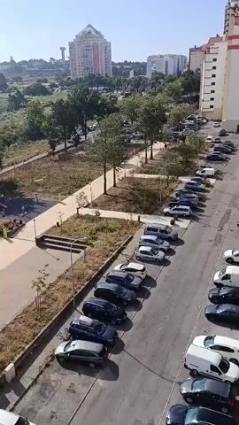 This media file captures a scenic view of Algueirão-Mem Martins, Portugal, on a bright, sunny day. The scene unfolds from a high vantage point, likely a balcony or window, overlooking a residential area and a public park.

In the foreground, a street is lined with parked cars, showcasing a variety of vehicles. Adjacent to the street is a modern-looking park with distinct areas for recreation. One section features a well-maintained skatepark with ramps and ledges. Another area is dedicated to sports, with a vibrant blue court that appears to be a basketball or futsal court, complete with hoops and markings. A portion of a tennis court with a blue surface and net is also visible. The park is surrounded by a fence and features paved pathways, benches, and lampposts, suggesting a well-designed public space. Grassy areas and scattered trees populate the park, blending it with the natural landscape.

Beyond the park, the view expands to reveal a residential neighborhood with buildings of varying heights, including a prominent tall building in the distance. A water tower is also visible on the horizon. The background is dominated by rolling hills covered in lush vegetation and a network of paths, indicating a semi-rural or suburban setting. The overall impression is one of a peaceful, sunny day in a well-equipped urban area bordering natural landscapes. No people are clearly visible in the park or streets, suggesting a quiet moment in time. The time of day appears to be morning or early afternoon, given the bright sunlight and shadows. The weather is clear and pleasant, ideal for outdoor activities. No discernible text is visible in the media.