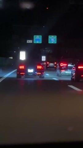 The video shows a car driving at night on a highway behind other cars. The car is following two other cars with red taillights. The road markings are visible and there are overhead highway signs indicating directions to "Bülach Flughafen" and "Bülach Kloten". The car is passing under the signs.