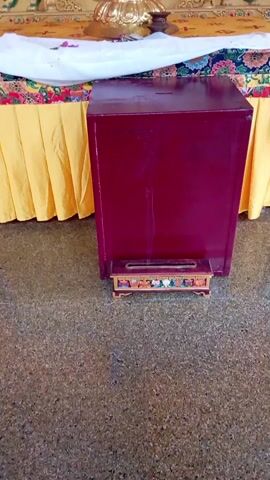 The camera pans up to reveal a burgundy rectangular donation box beneath an ornate golden Buddhist altar. The altar is draped with yellow and patterned fabric. A large golden bowl sits on top, filled with offerings. Above the bowl is a golden statue of Buddha. The background is covered in orange and gold decorations. The floor is speckled.