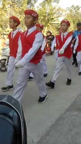 A group of young people, likely students, are dressed in matching uniforms and participating in what appears to be a drill or performance. The uniforms consist of white pants, white shirts, and red vests. Some individuals are also wearing red headbands or hats. They are standing in formation on an asphalt surface, possibly a road or a courtyard. In the background, there are trees and some structures. The camera is positioned at an angle, and the footage is rotated. The overall impression is of an organized activity involving a youth group.