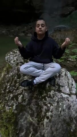 A person sits in a meditative pose on a rock in front of a waterfall. The person is wearing a black hooded sweatshirt, light-colored pants, and black shoes. They have a short haircut. The waterfall cascades down a rocky cliff face into a small pool of water. The surrounding environment is lush with greenery. The camera angle shifts upward to showcase the full height of the waterfall.