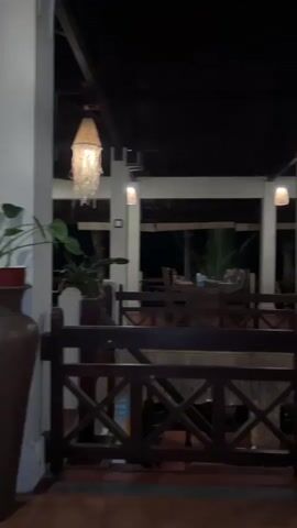 Here is a description of the video:

The video shows an outdoor space, likely a patio or deck, at night. The area is covered by a dark-colored roof, and supported by white columns. Wooden railings with an "X" pattern surround the space, creating a barrier. Large brown vases holding plants are placed around the perimeter. The floor is tiled in a reddish-brown color. Woven lamps hang from the ceiling, providing warm lighting. Through the openings in the railing, tables and chairs can be seen, suggesting a dining area. The scene appears to be a restaurant or resort.