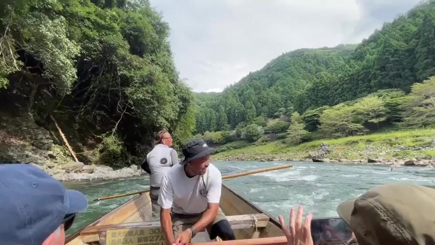 A group of people are on a boat in a river. The boat is being steered by a man in the front wearing a white shirt and a black hat, who is using two oars. Another man is standing behind him, also appearing to be part of the crew. In the foreground, there are parts of two other people visible from the chest up, one wearing a blue baseball cap and the other a beige hat, suggesting they are passengers. The river is wide and appears to have some rapids. The surrounding environment is lush and green, with steep, tree-covered hillsides flanking the river. The sky is partly cloudy. The overall impression is a scenic boat tour through a natural landscape.
