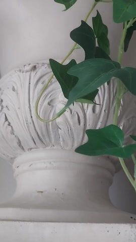 A close up video of a white vase with ornate carvings on it. The vase is filled with artificial plants. The plants are green ivy and a large light green hydrangea flower. Some thin white branches are also sticking out of the vase. The video is shot from a close up, vertical angle.