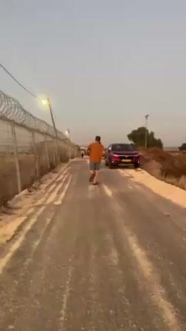 The video shows a road beside a fenced area with razor wire. There are people, a red car, and several evenly spaced lights. The sky is light blue. The road is paved with dirt and tire tracks.