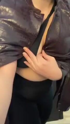 The video shows a woman wearing a black bra, black leggings, sunglasses and a brown jacket. She is moving erratically and adjusting the jacket in front of the camera. Behind her are a brown door and an easel with a colorful painting. A partial view of the room is visible through the open doorway. Loud music is playing in the background.