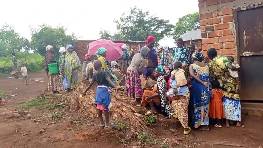 A group of people, mostly women and children, are gathered outdoors. Most of the women are wearing colorful patterned dresses and head coverings. Some are carrying children. One woman holds a pink umbrella. A child holds up a small yellow object. Some people are standing while others are seated on plastic chairs. The ground is muddy. In the background, there are buildings with brick walls. The environment appears to be rural, with trees and vegetation visible.