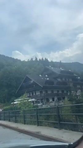 The video shows a scenic drive through Scopello, Italy, with a focus on the natural beauty and architecture of the region. The scene opens with a large, multi-story building with a dark, pitched roof, characteristic of mountain architecture, nestled amongst lush green trees. The building has multiple balconies and appears to be a hotel or resort, with white umbrellas suggesting an outdoor dining area.

As the camera pans, the landscape transitions to reveal a picturesque mountain range in the distance, shrouded in a hazy, overcast sky. A river flows through the valley, its rocky bed and rushing water adding to the natural charm. Another building, smaller and lighter in color, is visible on the hillside, also integrated with the natural environment. The video captures the essence of a serene mountain town, with a blend of man-made structures and the raw beauty of nature. The weather appears to be cloudy, perhaps indicating an early morning or late afternoon, with the diffused light softening the overall scene. The overall impression is one of peacefulness and natural splendor, inviting viewers to experience the tranquility of Scopello.