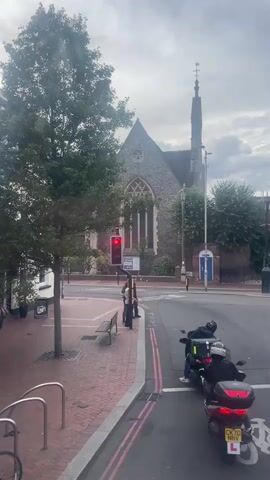 The video opens on a street scene with a prominent church in the background. A traffic light is red, and two individuals are waiting on the sidewalk next to a bench. A tree stands to the left of the frame, and to the right, two people on scooters are stopped at the intersection. The scooter in the foreground has an L-plate. A cyclist then rides past the stationary scooters and the church. The sky is overcast.
