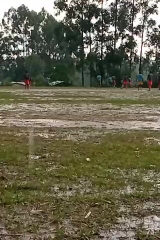 The video shows a group of people playing soccer on a muddy field. The field is wet and slippery, with puddles of water visible. There are trees in the background. The players are wearing different colored shirts. They are running around the field, kicking and passing the ball. The video is shot from a distance.