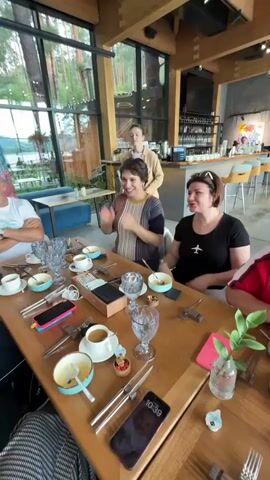 The video shows a group of people sitting at a long wooden table in what appears to be a restaurant or cafe. The table is set with plates, cups, and silverware, indicating a meal or celebration. Several people are clapping, and one woman in a yellow jacket is handing a gift bag to another woman at the table. The environment seems to be indoors with large windows offering a view of trees and possibly water. There is a bar area in the background. The overall mood appears festive.