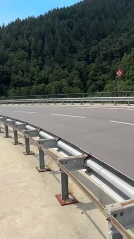This is a vertical video clip filmed from a moving vehicle, likely a car, on a highway. The scene depicts a scenic view of a dense, green forest covering a steep hillside. The road itself is paved and has white lane markings. A silver metal guardrail runs along the side of the road. In the distance, a red circular speed limit sign with "50" in white is visible on a post.

The sky is a bright blue with scattered white, fluffy clouds, suggesting fair weather. The sun appears to be high in the sky, indicating it's likely daytime, perhaps late morning or early afternoon. The overall mood conveyed is one of travel and appreciation of nature, with the lush greenery and open sky creating a serene atmosphere.

Further in the distance, the road curves, and a bridge or overpass can be seen. Below this, a set of train tracks is visible, hinting at the transportation infrastructure in the area. The foliage is rich and varied, with different shades of green indicating a healthy, thriving ecosystem. The video captures the essence of a road trip through a mountainous, forested region, likely in Germany given the location context of Forbach. There are no people visible in the clip.