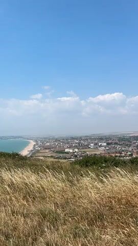 A high-angle, wide shot shows a coastal town seen from a grassy hilltop. The sky is bright blue with scattered white clouds. In the foreground, tall, dry grass sways in the wind. Below, the town stretches along a wide, sandy beach bordering the turquoise ocean. Buildings of varying heights, mostly red-tiled roofs, fill the urban area. The scene is bathed in natural light, suggesting a clear, sunny day. There are no people visible in the frame.