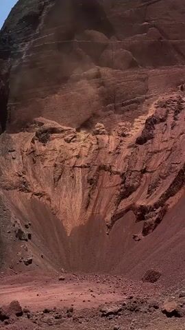 The video shows a steep, rocky landscape, possibly a crater or canyon, with reddish-brown rock formations and loose gravel. A plume of dust or smoke is rising from the upper right portion of the scene, indicating a disturbance or ongoing geological event. The lighting suggests it is daytime.