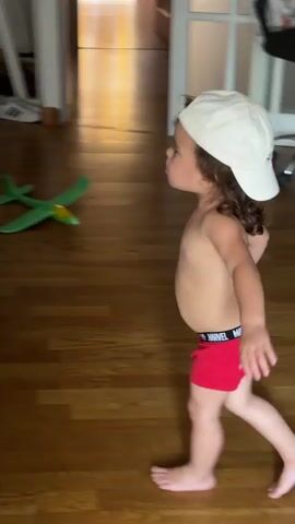 Two young children are in a living room, a common domestic setting. One child, wearing a white baseball cap and red Marvel-branded underwear, is in the foreground, facing away from the camera and towards the other child. Their arms are extended, as if reaching out or dancing. The second child, seen in profile and facing the camera, is leaning against a dark leather couch. They are wearing a diaper. Both children appear to be toddlers.

The room has wooden flooring and a dark leather couch is a prominent piece of furniture. A green toy airplane lies on the floor behind the children, suggesting a playroom or family area. In the background, a doorway leads to another part of the house, where natural light suggests it is daytime.

The children's interaction is unclear, but the child in the foreground seems playful, while the child leaning on the couch has a more serious expression. The scene is indoors, and the lighting suggests it is daytime. There are no immediate clues about the weather.

Visible text includes "MARVEL" on the waistband of the red underwear and "LITTLE STEPS BIG PROGRESS" on the back of the white baseball cap. The location is identified as Platja D'aro, Spain.