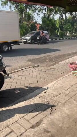 A white van with pink accents drives on the left side of a paved road. The van has advertising on its side. A gray truck with an open bed drives down the same road. Two people on a motorcycle drive past a silver SUV on the road. The camera is focused on a brick sidewalk.
