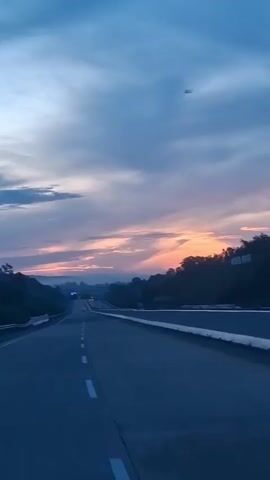 The video shows a view of a highway from a moving vehicle at dusk. The sky is a deep blue with scattered clouds that are tinged with pink and orange from the setting sun. The highway is empty except for a few distant vehicles, and it stretches into the horizon. The surrounding landscape is dark and hilly, covered in trees. The camera is angled slightly upwards, capturing the vastness of the sky and the open road.