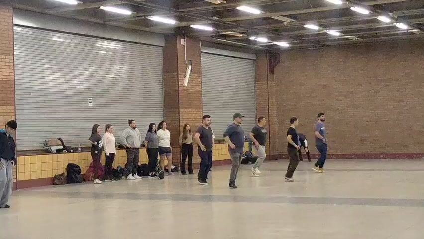 A group of approximately 15 people are gathered in a large, indoor space with a polished concrete floor and brick walls. The space appears to be a gymnasium or multipurpose room, with large, corrugated metal doors visible in the background. Overhead, fluorescent lights illuminate the area. Most of the people are standing in a loosely formed circle, with several individuals appearing to be dancing or practicing movements. One person on the far left is actively engaged in a dance move, while others observe or participate in a less energetic manner. Bags and other personal belongings are scattered on the floor near the wall on the left. The overall atmosphere suggests a casual rehearsal or informal dance session.