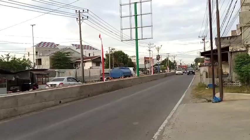 The video shows a street scene with traffic. On the left side of the road, there is a concrete barrier with some people sitting near it. A white car is parked behind the barrier. Further down the street, there are buildings, a billboard, and power lines. Several vehicles, including cars, a truck, and a motorcycle, are driving on the road. The sky is cloudy.