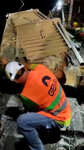 The video shows construction workers at night. A person in an orange safety vest and white hard hat is crouched down, reaching into a pile of debris. Another person wearing a yellow hard hat is visible in the background, also appearing to be involved in the work. The scene is dark, with artificial lighting illuminating the area. There are broken structures and pipes among the debris, suggesting a collapse or demolition. The overall environment appears to be a construction site or an area affected by structural damage.