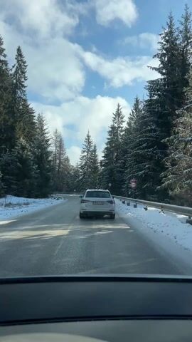 The video shows a view from inside a vehicle driving on a snowy road through a forest. The road is covered in slush and appears slick. The forest consists of tall, coniferous trees heavily laden with snow, lining both sides of the road. A white car is driving ahead, maintaining a safe distance. The sky is blue with scattered white clouds. There are road signs visible, including one indicating a speed limit of 40. The camera is steady, providing a clear view of the winter scenery.