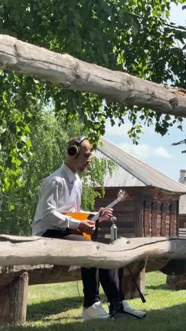 A young man wearing headphones and a white button-down shirt sits on a wooden bench outdoors and plays a balalaika. He is positioned in front of a microphone, and appears to be singing or speaking into it as he plays. The background features greenery, trees, and a wooden building with intricate carvings. A rough-hewn wooden fence runs across the foreground of the frame.
