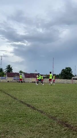 A group of men wearing yellow vests and red shorts are practicing soccer on a grassy field. The sky is cloudy. There are buildings and trees in the background.