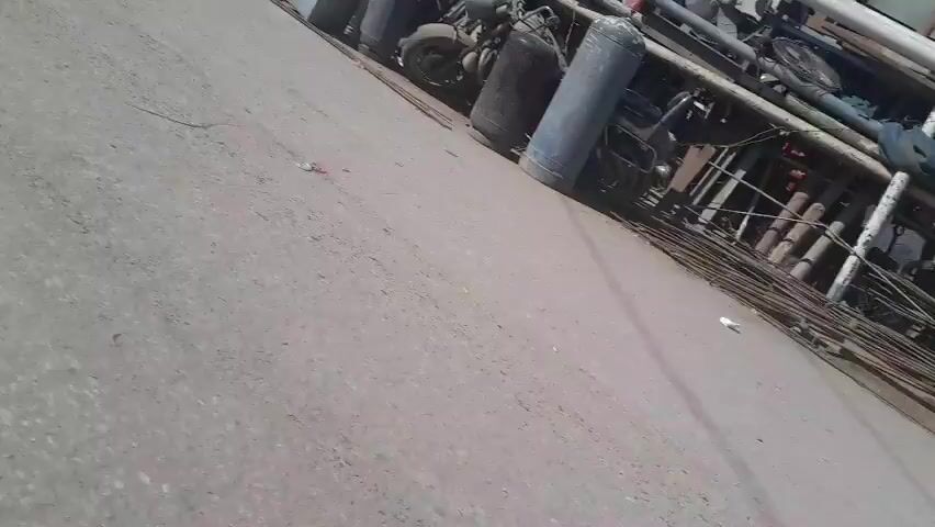 The video shows an outdoor scene with a concrete surface in the foreground and a scrap metal yard in the background. The scrap yard contains various items including metal pipes, gas cylinders, what appear to be construction materials, and a blue plastic barrel. A white vehicle is visible in the distance. The sky is clear and blue, suggesting it is daytime. The camera appears to be moving, likely handheld.