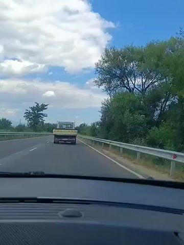 A video shows a view from inside a car traveling on a highway. The car is moving along a paved road with a white line marking the lane. To the right of the road, there is a metal guardrail. Beyond the guardrail and the road, there are green trees and bushes lining the scenery. In the distance, a white truck with a yellow bed is driving in the same direction as the car, ahead on the highway. The sky above is blue with scattered white clouds. The video appears to have been recorded during the daytime.