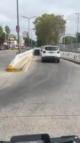 From a low angle, a white Jeep driving away from the camera moves into an underpass. The walls on either side are decorated with colorful triangles. Street lamps line the sides of the road. The road is made of gray concrete. There is light traffic on the street parallel to the road on top of the underpass. The sky is overcast.
