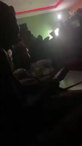 The video shows a dim room, possibly a lounge or bar, with people sitting on couches and chairs around tables. The room is illuminated with ambient blue and purple lighting. Several individuals are visible, though their features are indistinct due to the low light. They appear to be engaged in conversation and using their phones. A window with bars is visible in the background. The overall atmosphere seems relaxed and intimate.