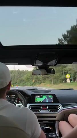 The video shows the interior of a car with a panoramic sunroof open. The camera is positioned behind the driver and a passenger, looking forward through the windshield and sunroof. The driver, wearing a white baseball cap and a light-colored polo shirt, is visible from the back. The passenger's legs are visible in the lower right of the frame. The car's dashboard is illuminated, with a navigation screen displaying a map and route. The car is driving on a paved road through a wooded area with green trees and foliage on either side. There are yellow warning signs visible along the road. The sky above is bright and appears overcast.