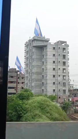 This video shows a street view from inside a vehicle. The first shot shows a tall, unfinished concrete building with an Argentinian flag on its roof. Several flags can be seen on other buildings in the background, including a green flag with white writing and what appears to be an Argentinian flag. The video pans across a cityscape with various buildings of different heights and architectural styles. There are power lines and utility poles throughout the scene. In the foreground, there is a concrete barrier with yellow and black markings. The sky is overcast.