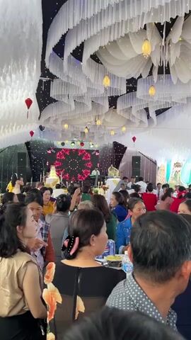 This video captures a lively wedding reception or large social gathering held under a decorated tent in Bình An, Vietnam. Numerous guests, predominantly adults of various ages, are seated at round tables covered with white tablecloths, engaged in conversation and observing the event.

The setting is elaborately decorated, featuring a ceiling adorned with intricate white draped fabric, interspersed with warm fairy lights and hanging lanterns (both red and yellow/gold). A stage is visible at the far end, featuring a large circular LED screen displaying a vibrant red geometric pattern and a multi-tiered wedding cake. Speakers and other event equipment flank the stage.

Guests are dressed in casual to semi-formal attire, typical for such celebrations. Fans are suspended from the ceiling, suggesting warm weather. Natural light, likely from the tent's open sides, indicates it is daytime. A Pepsi can is visible on one of the tables. The overall atmosphere appears festive and engaging, with people enjoying the celebration and the company of others. In the background, glimpses of greenery and what appear to be residential structures can be seen.