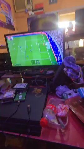 A video shows a person playing a soccer game on a PlayStation 4. The game is being played on a Samsung TV. On the table in front of the TV are a PS4 console, several external hard drives, two Playstation controllers, some rubber bands and a plastic bottle with a red cap. The room has brown walls and what appears to be a poster with “Accessories” on it is partially visible. An air conditioner is on the wall. The video is shaky and appears to be taken from a first person perspective.