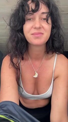 A woman with long, dark, curly hair is shown from the chest up in a bedroom. She is wearing a grey sports bra and black leggings. She is in the process of taking off a grey long-sleeved shirt with lime green accents. She pulls the shirt over her head, her hair falls forward, and she shakes her head, causing her hair to spread out. She is smiling and looking at the camera. The background consists of wood-paneled walls and a bed.