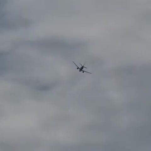 An airplane is visible in the sky, appearing as a small silhouette against a cloudy background. The airplane is in mid-flight, with its wings and tail clearly discernible. The clouds are dense and overcast, obscuring much of the sky. The camera is pointed upwards, capturing the airplane from below. The lighting is dim, consistent with an overcast day. There are no other visible objects or people in the frame.