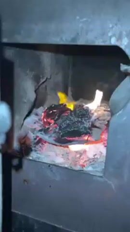 The video shows a small fire burning inside a metal fireplace or stove. The fire consists of small flames and glowing embers over a bed of ashes. The interior of the fireplace is dark and sooty, with visible wear and tear on the metal surfaces. A portion of an arm or sleeve is visible on the left side of the frame, but no person is fully visible. The camera is relatively steady while the fire burns.