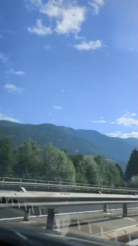 The video shows a view from inside a vehicle moving along a highway. A bright blue sky with scattered white clouds is visible above a range of green mountains. The mountains are covered with dense trees, primarily evergreens. A guardrail runs along the side of the highway in the foreground, and a white car is seen driving in the same direction. The camera appears to be looking slightly upwards towards the sky and mountains.
