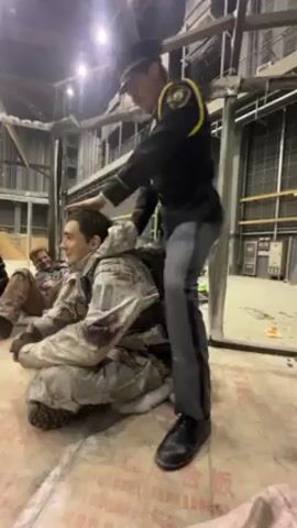 A man in a police uniform is standing over a man in a camouflage uniform who is sitting on the floor. The man on the floor is covered in what appears to be blood or fake blood. The police officer has his hands on the man's back and appears to be restraining him. Another man is lying on the floor in the background. The scene appears to be a movie or TV set, as there are metal frames and unfinished walls visible. The police officer is wearing a black uniform with yellow accents, and the man on the floor is wearing a camouflage uniform with bloodstains. The lighting is bright and artificial.